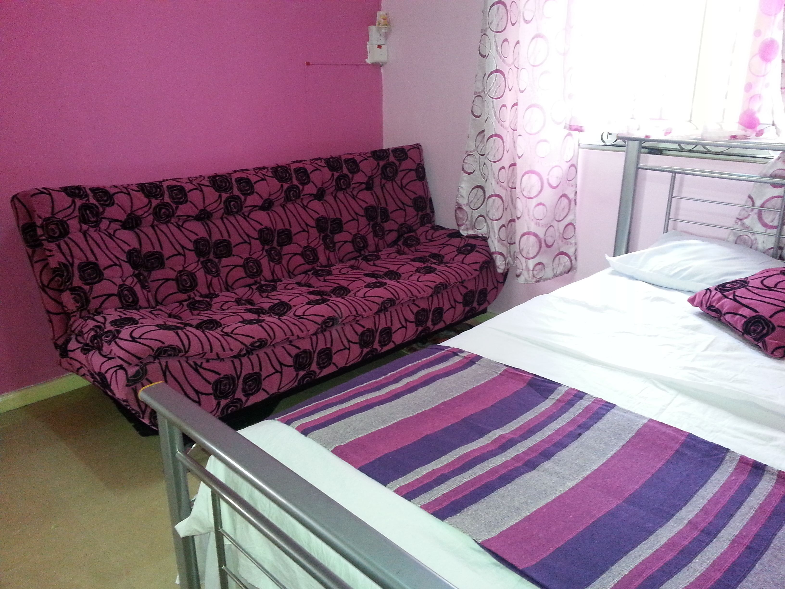 A bedroom with a pink wall, a bed with white bedding, a striped rug, and a pink floral sofa.