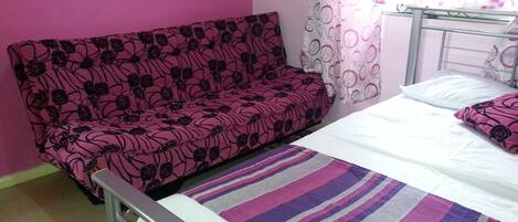 A bedroom with a pink wall, a bed with white bedding, a striped rug, and a pink floral sofa.