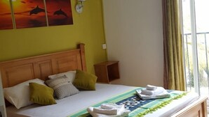 A hotel room with a large bed, a ceiling fan, an air conditioning unit, a window with curtains, and a wall with a sunset painting.