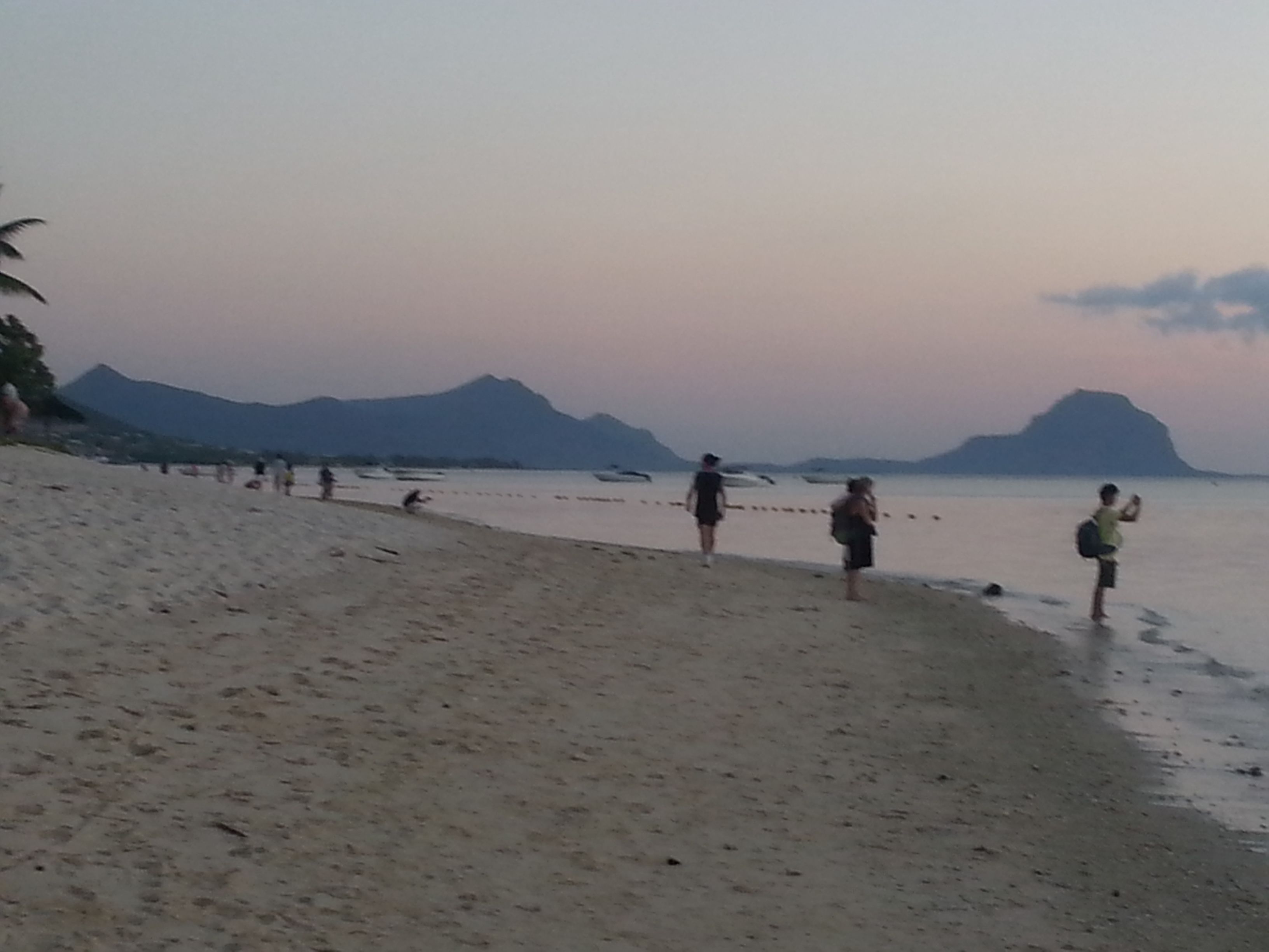 En strand med människor som promenerar längs strandkanten, ett lugnt hav och berg i bakgrunden.