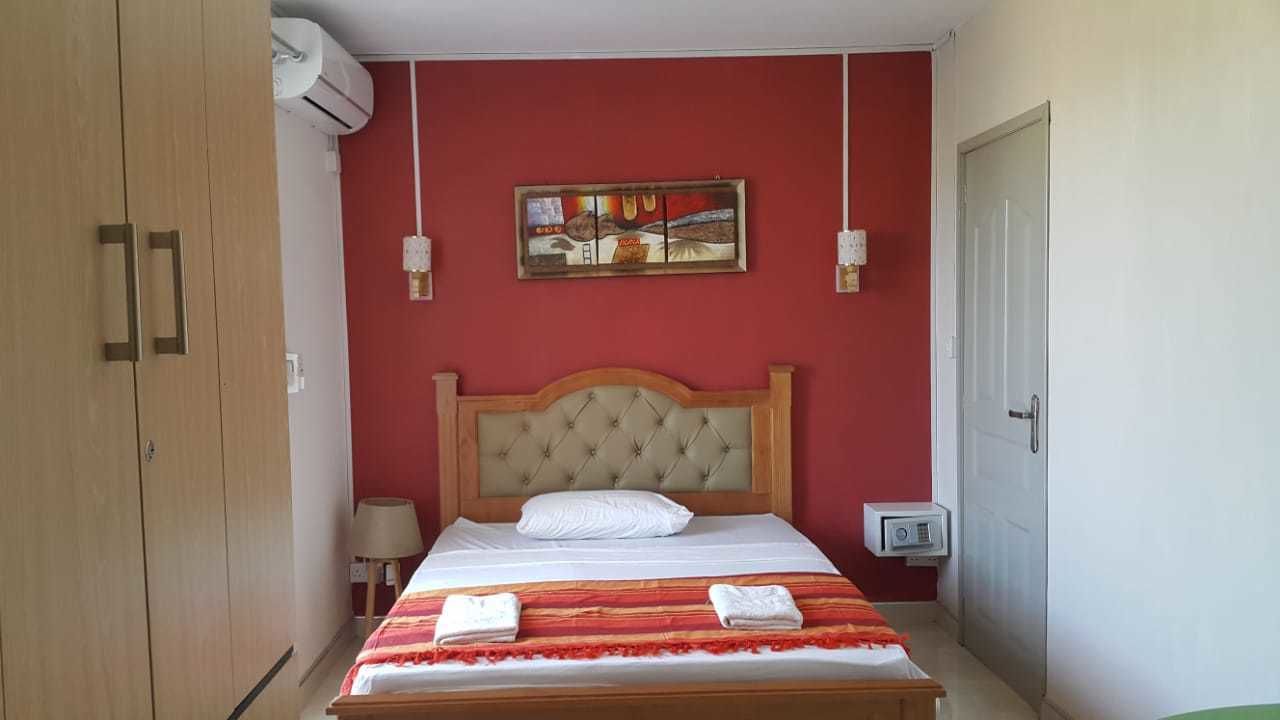 A bedroom with a wooden wardrobe, a bed with a tufted headboard, a red wall, and a painting above the bed.