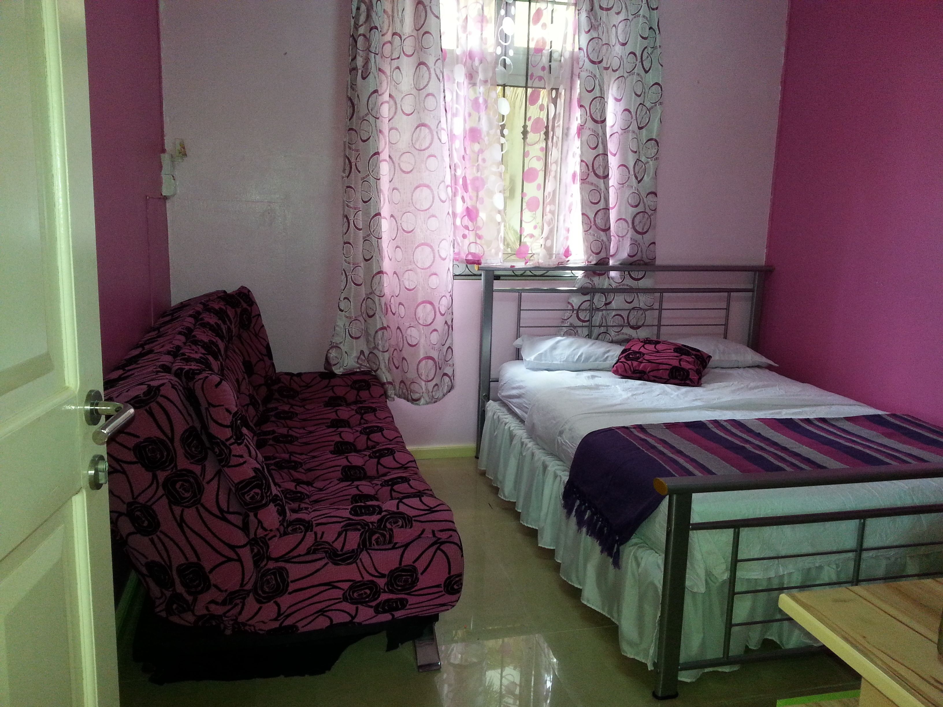 A small, single-bed room with a metal frame bed, a single sofa with a floral pattern, and a window with patterned curtains.
