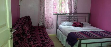 A small, single-bed room with a metal frame bed, a single sofa with a floral pattern, and a window with patterned curtains.