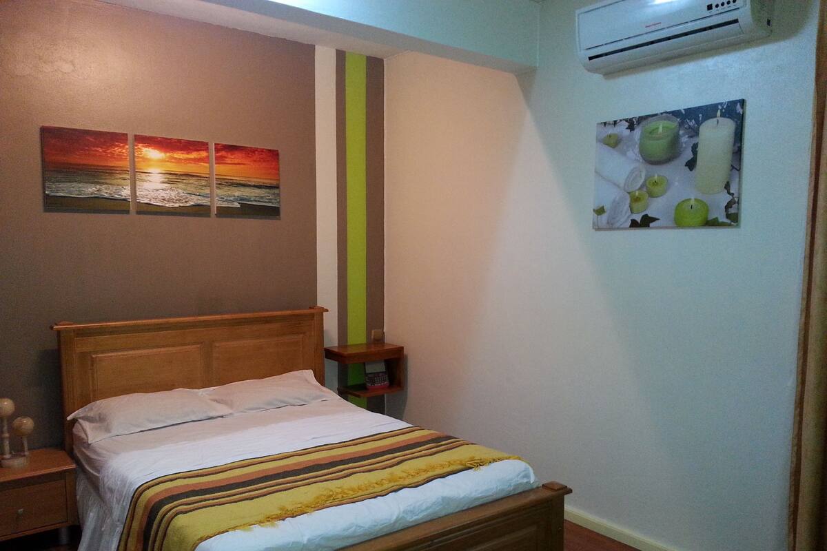 A bedroom with a wooden bed, a nightstand, a wall-mounted air conditioner, and two paintings.