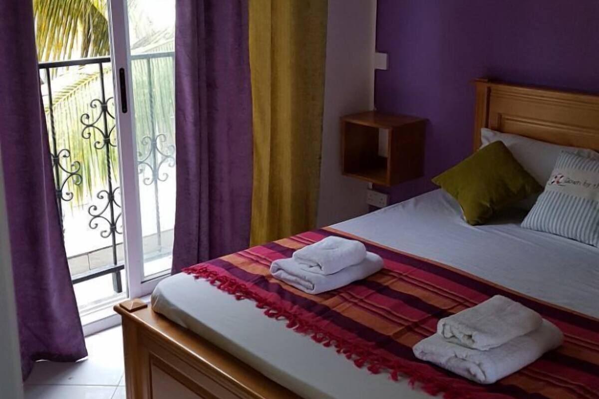 A bedroom with a bed, a nightstand, a wall-mounted lamp, and a view of the outdoors through a sliding door.
