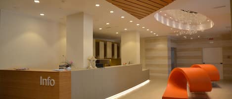 A reception area with a wooden reception desk labeled "Info," a modern chandelier, and orange seating.