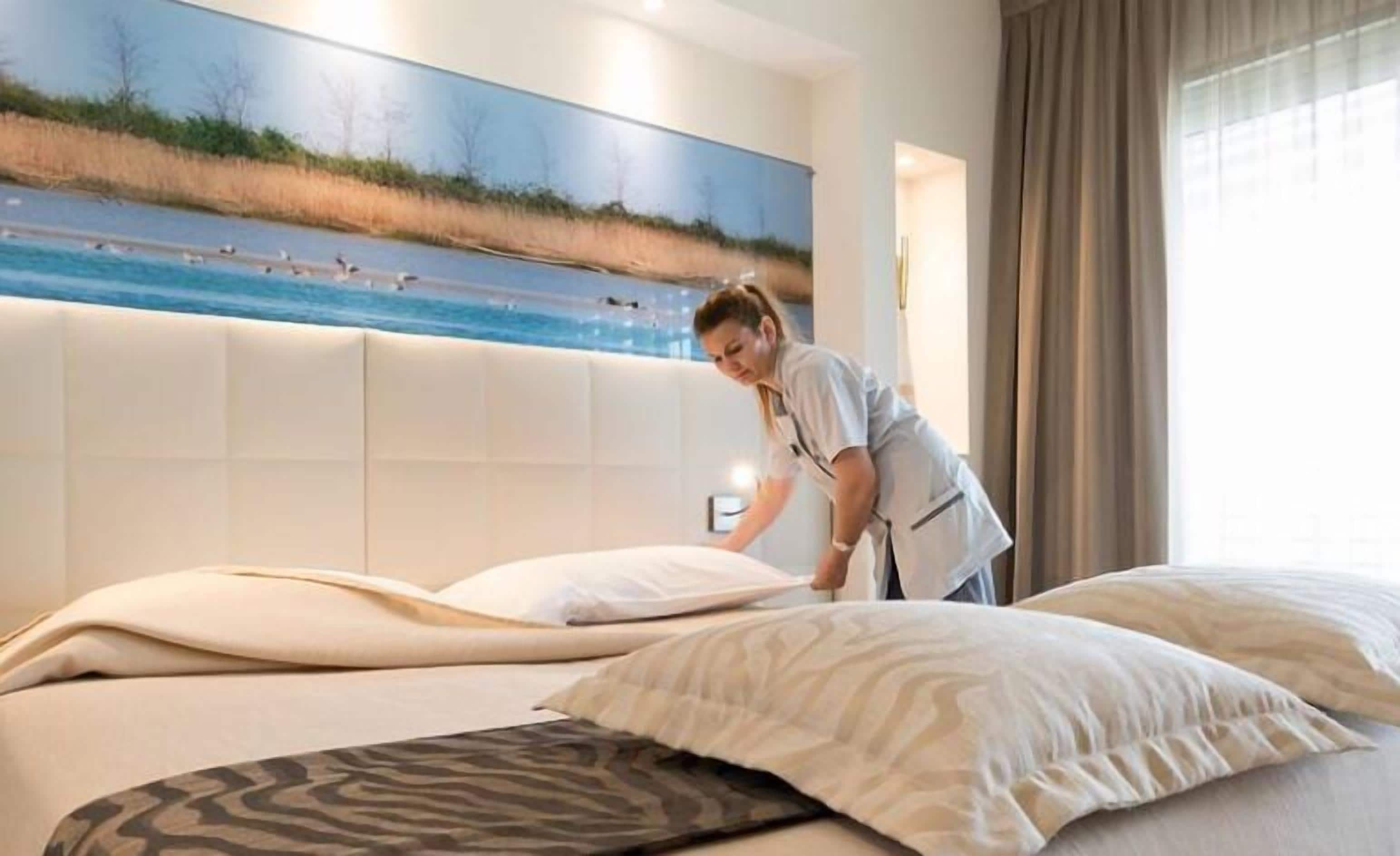 Een hotelkamer met een groot bed, een hoofdbord met een schilderij van een strandlandschap en uitzicht op zee.