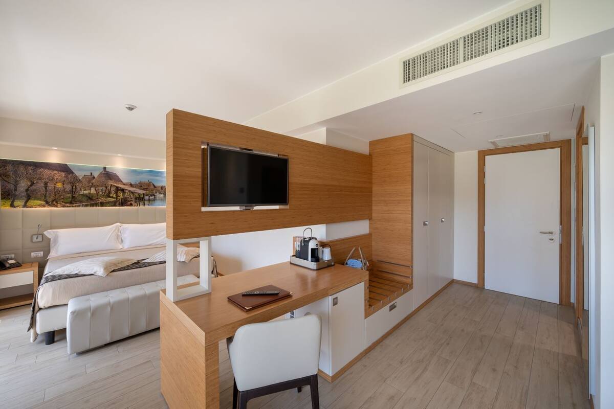 Een moderne hotelkamer met een bed, een bureau met een televisie, een koffiezetapparaat en een schilderij van een dorpsgezicht.