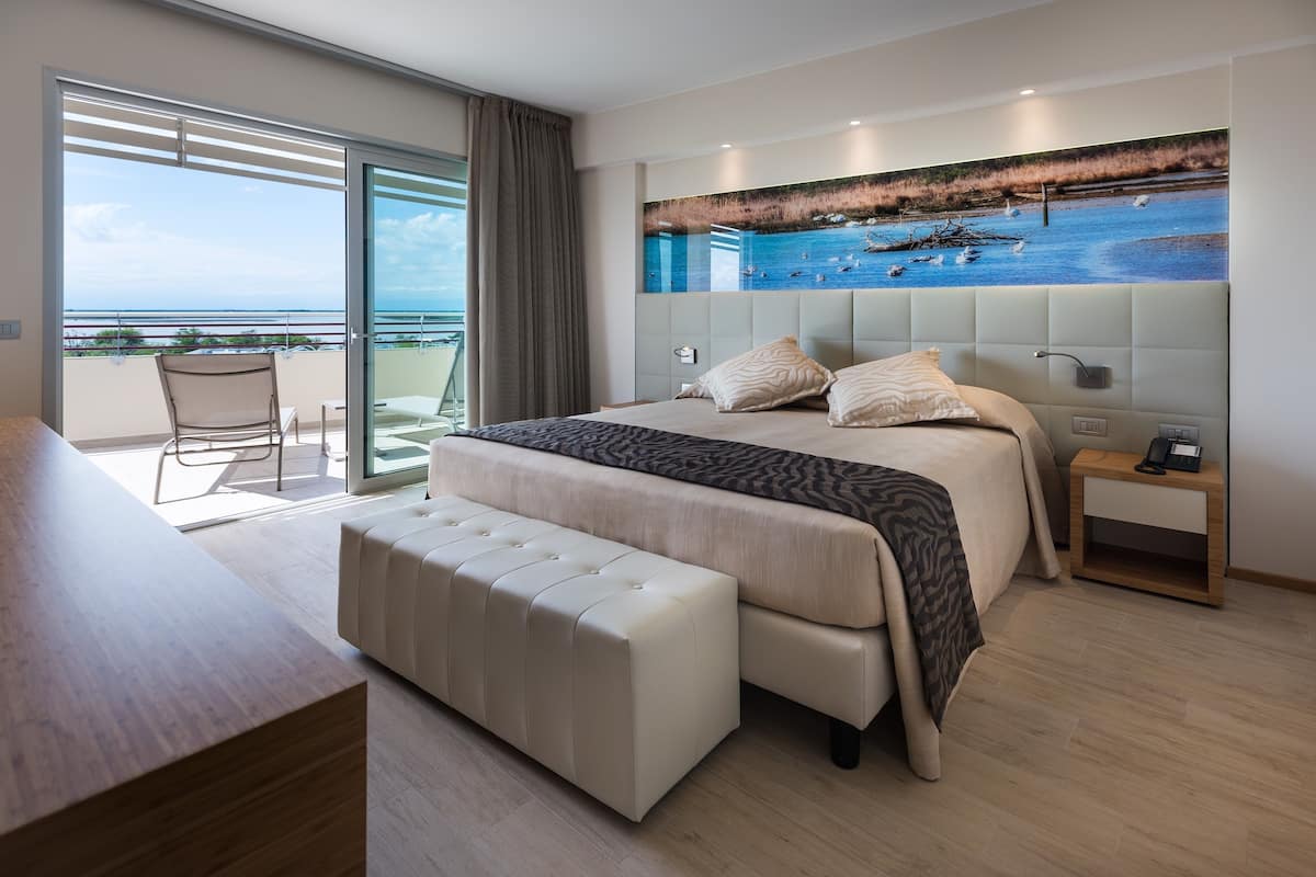 Een moderne slaapkamer met een groot bed, een bankje, een nachtkastje en uitzicht op zee door een schuifdeur.