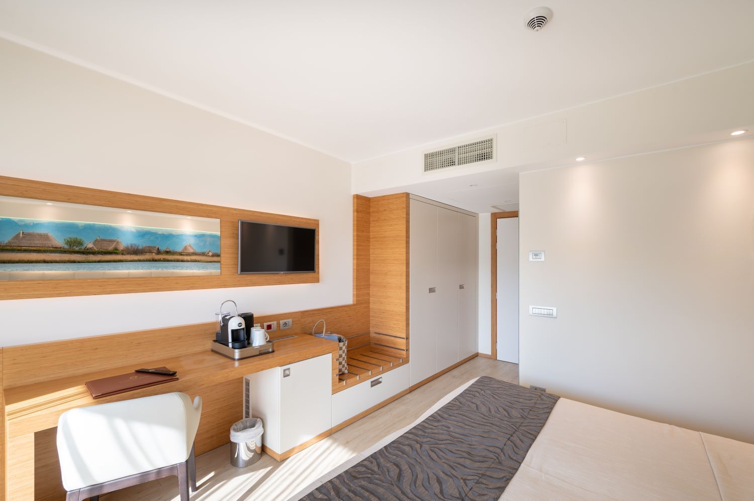 A modern hotel room with a wooden desk, a flat-screen TV, a coffee maker, and a large window.