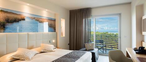A modern hotel room with a large bed, a desk, and a balcony with a view of the sea.