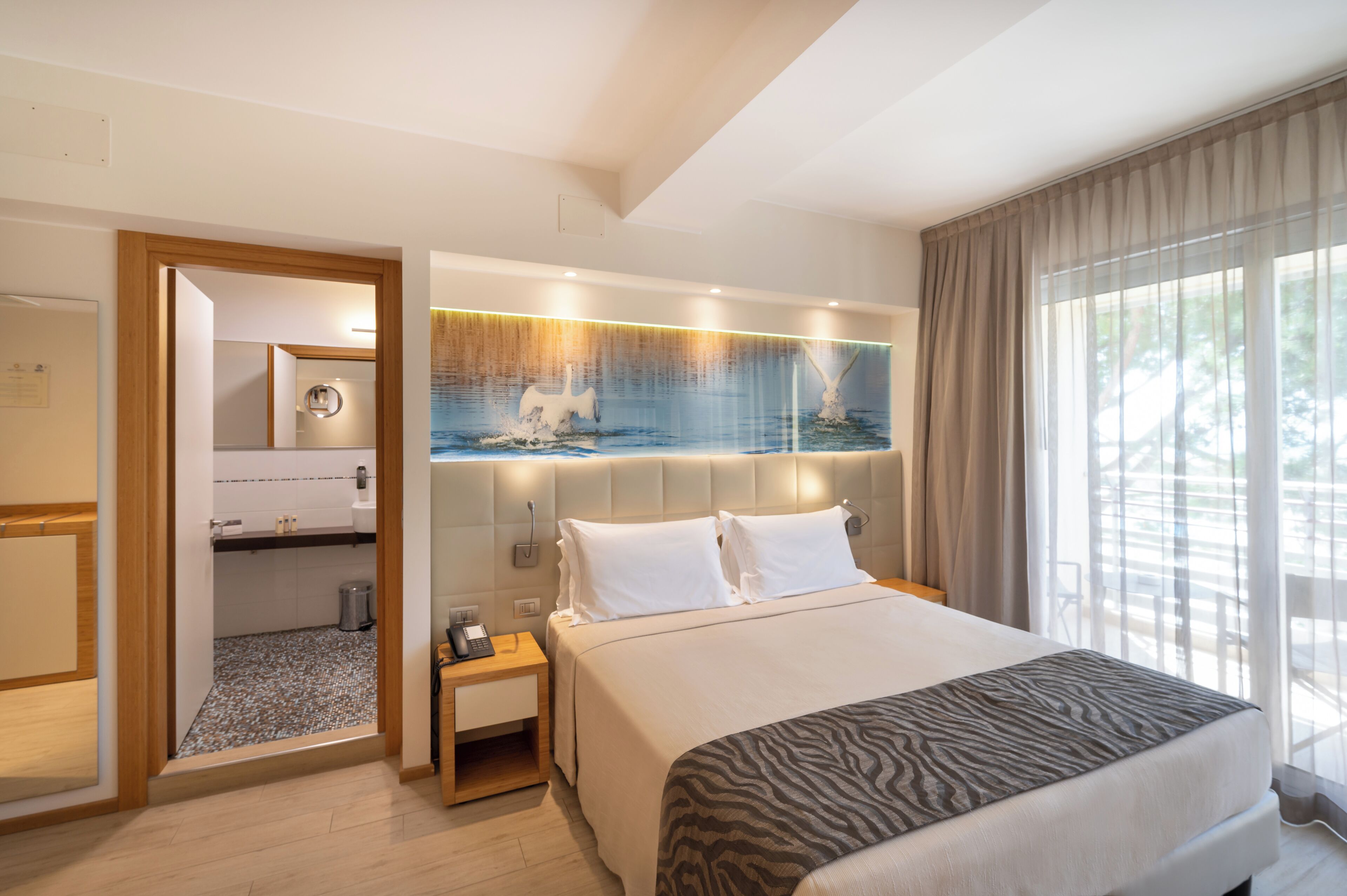 A hotel room with a large bed, a headboard with a decorative panel, a bedside table with a lamp, and a view of the outdoors through a window.