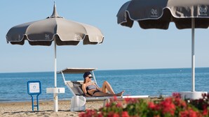 Eine Strandansicht mit einer Frau, die unter einem Strandsonnenschirm entspannt, einem Schild mit der Aufschrift 'AGUNA PARK' und freiem Blick auf das Meer.