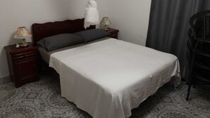 Una cama individual con una colcha blanca, dos mesitas de noche con lámparas, un cabecero de madera oscura y una cortina gris, además de una silla de madera oscura.