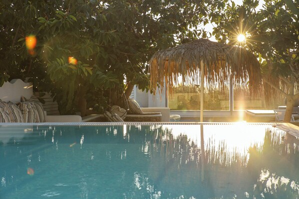 A swimming pool with a thatched roof structure, surrounded by lush greenery and a clear view of the sunset.