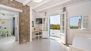 A room with a stone wall, a white bed, a desk with a chair, and a TV on the wall. - La Stella Stay (Mykonos)