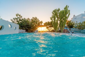 A pool with clear blue water, surrounded by white walls with circular cutouts, a large cactus, and a building with a dome in the background.