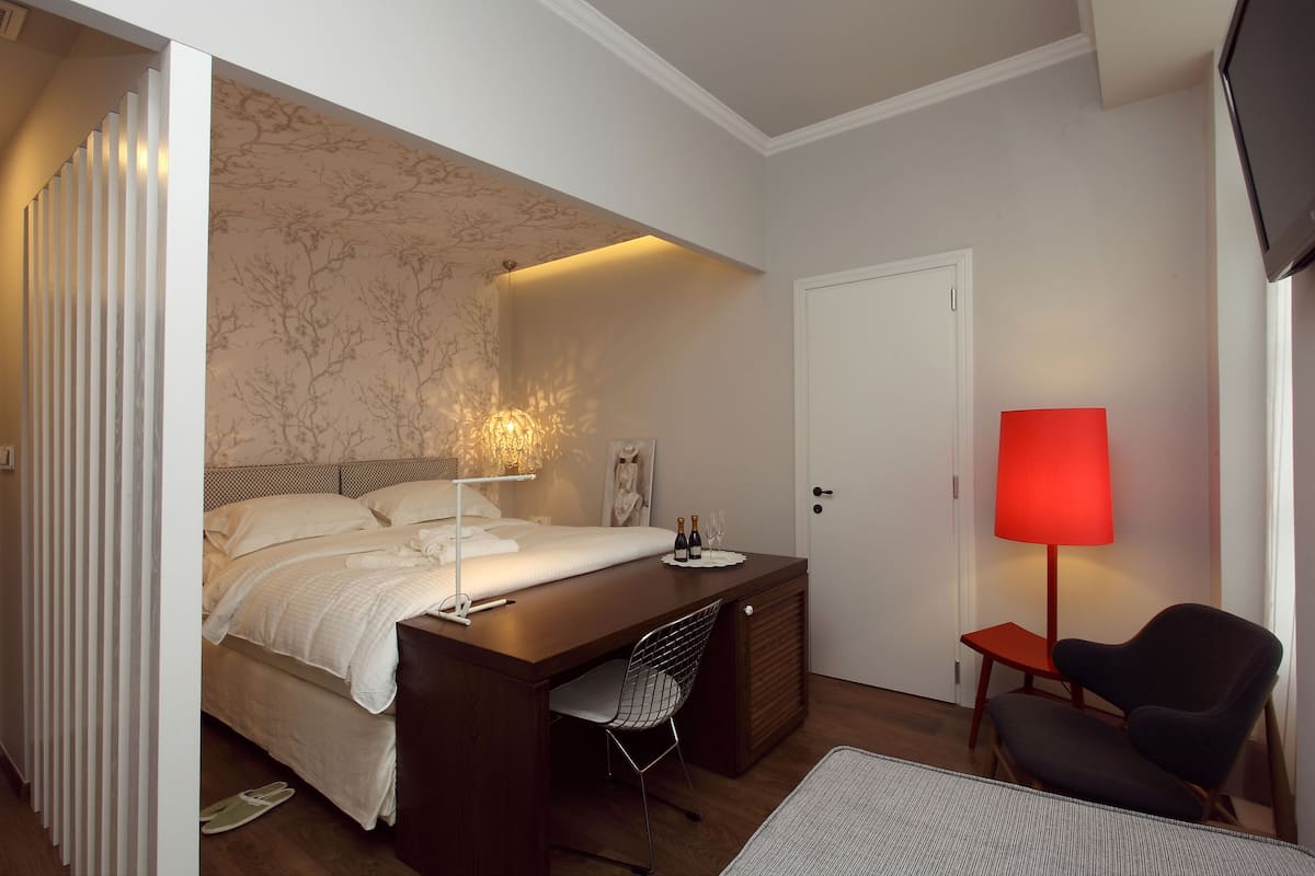 A bedroom with a bed, a desk, a chair, a red lamp, and a door.