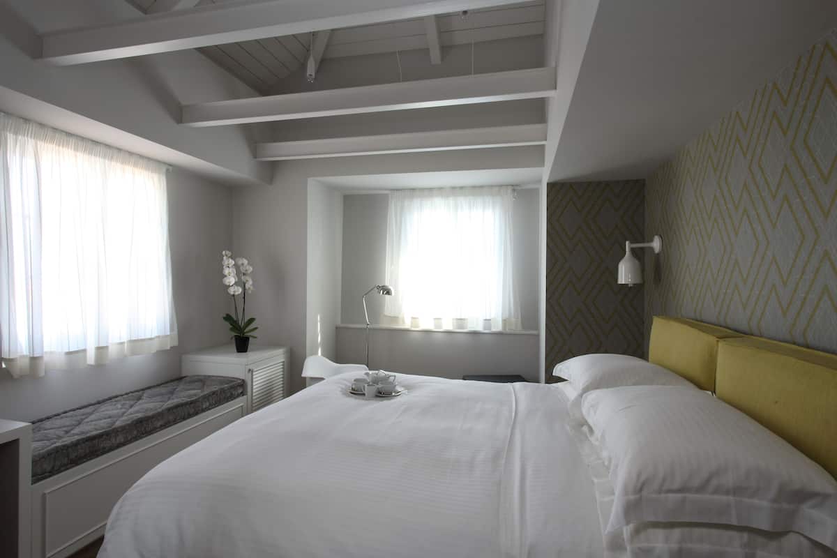 A hotel room with a large bed, a bedside table with a lamp, a window with curtains, and a decorative wall pattern.