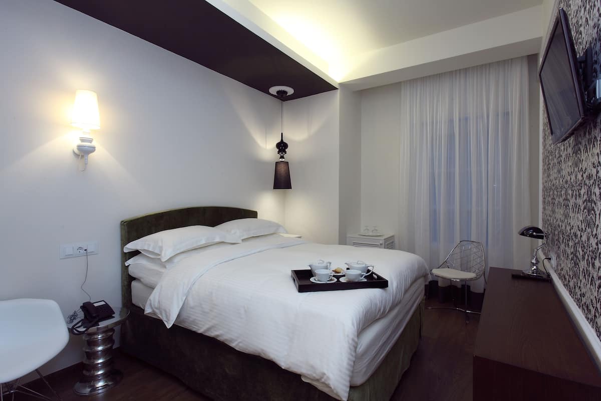 A hotel room with a bed, bedside table, chair, desk, and a tray with cups and a bowl.