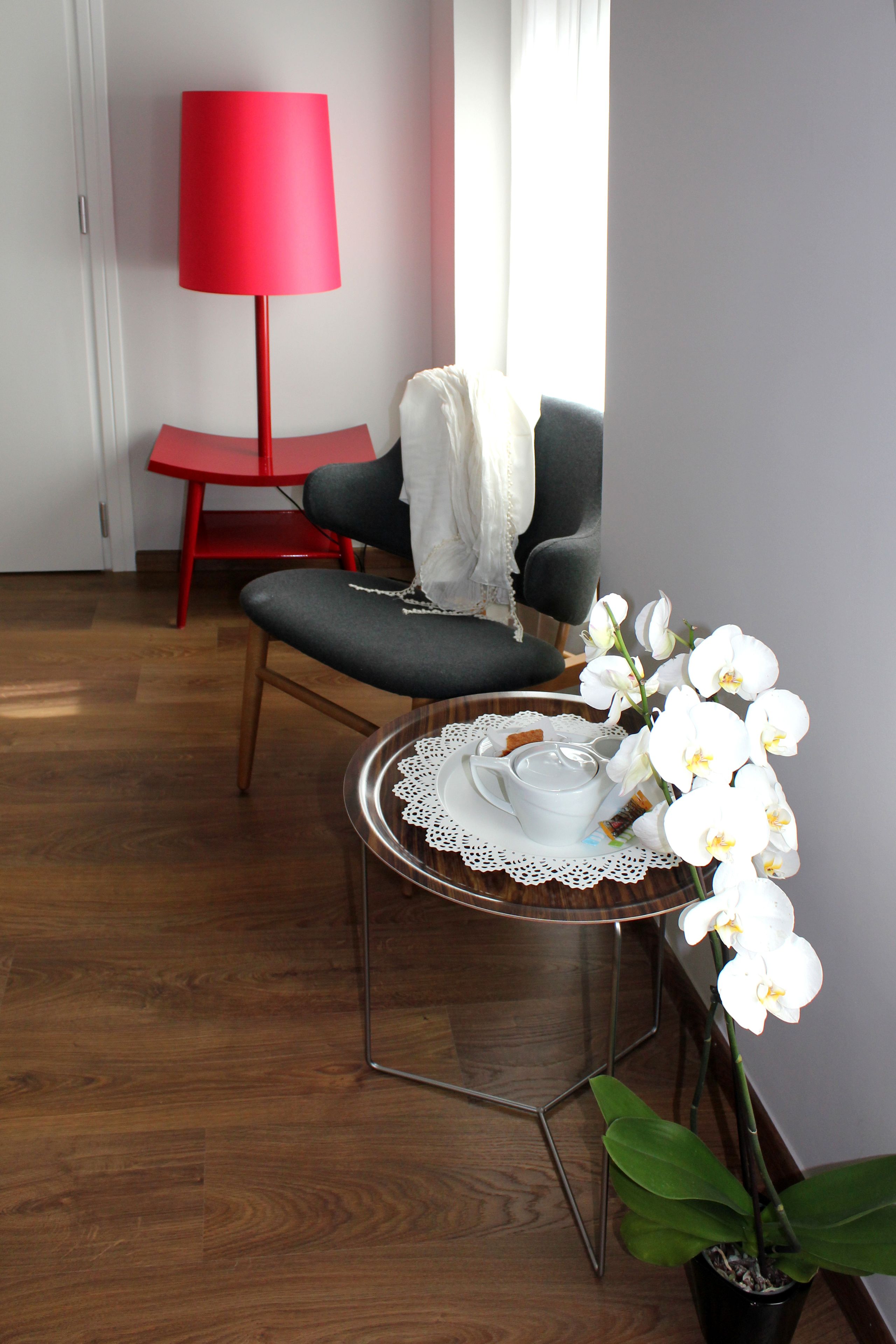A room with a wooden floor, a red chair, a small round table with a white cup and saucer, a white orchid plant, and a red floor lamp.