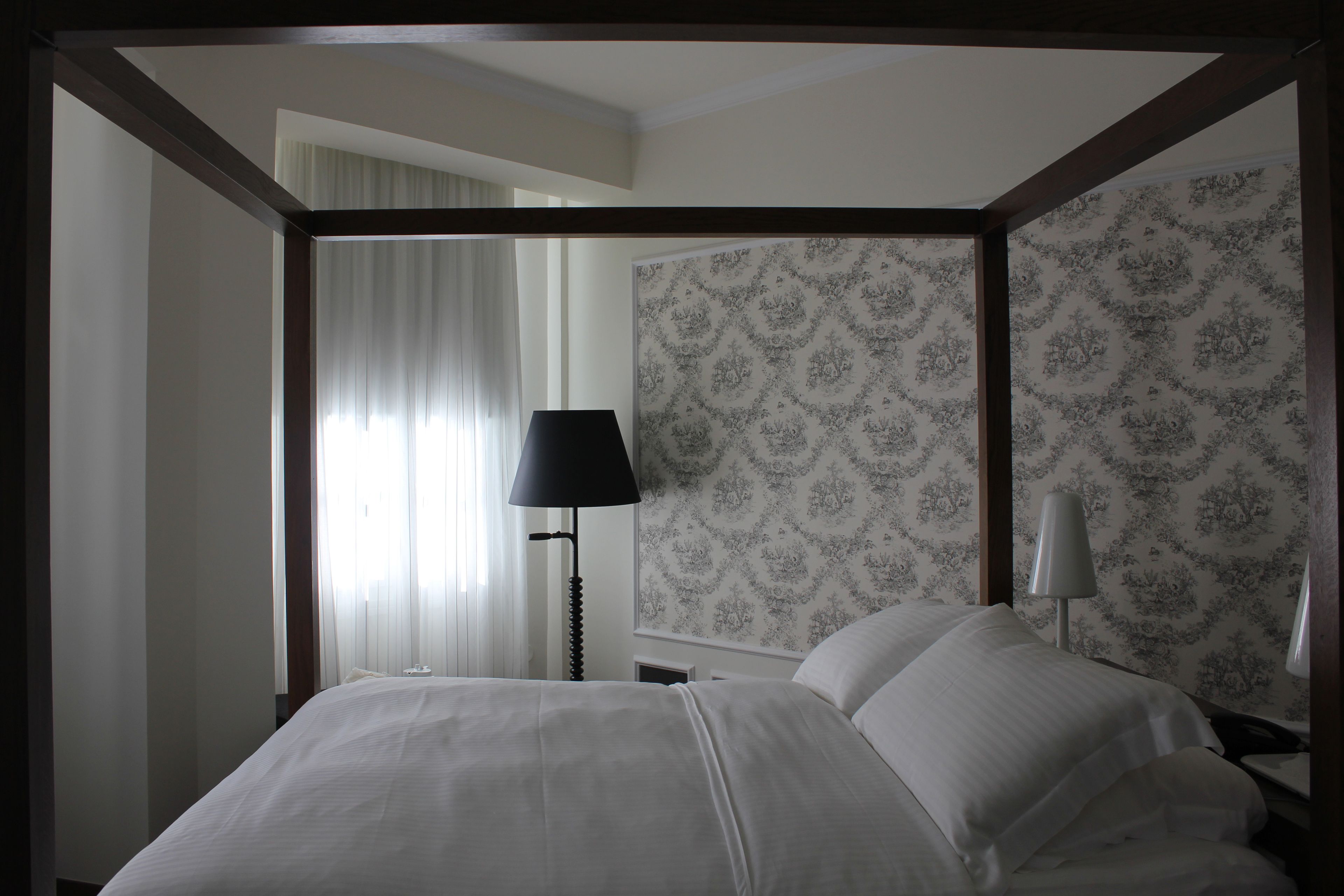 A hotel room with a large bed, a nightstand, a lamp, and a patterned wallpaper.