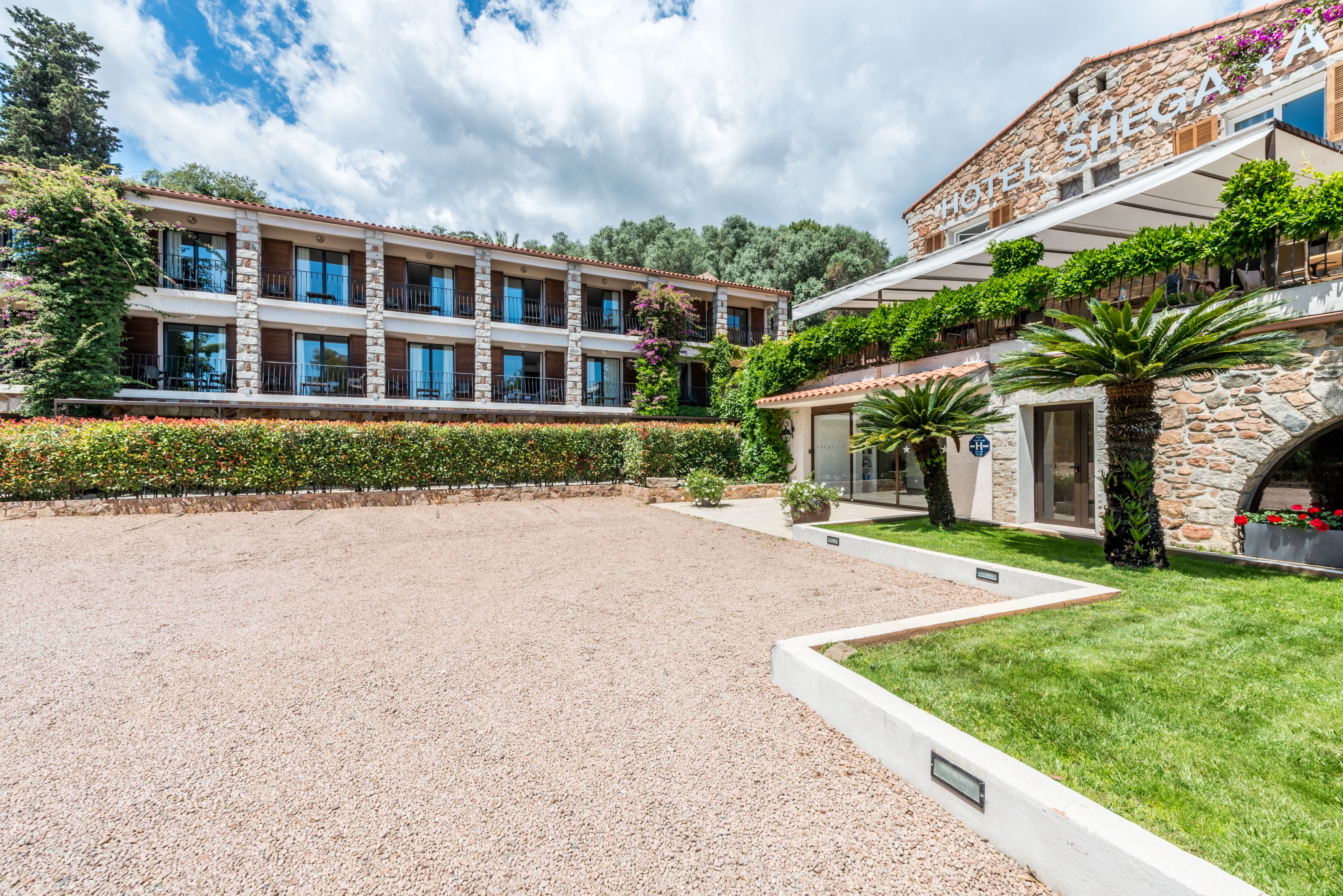 Um hotel com várias andares, fachada de pedra, varandas e um pátio de cascalho.