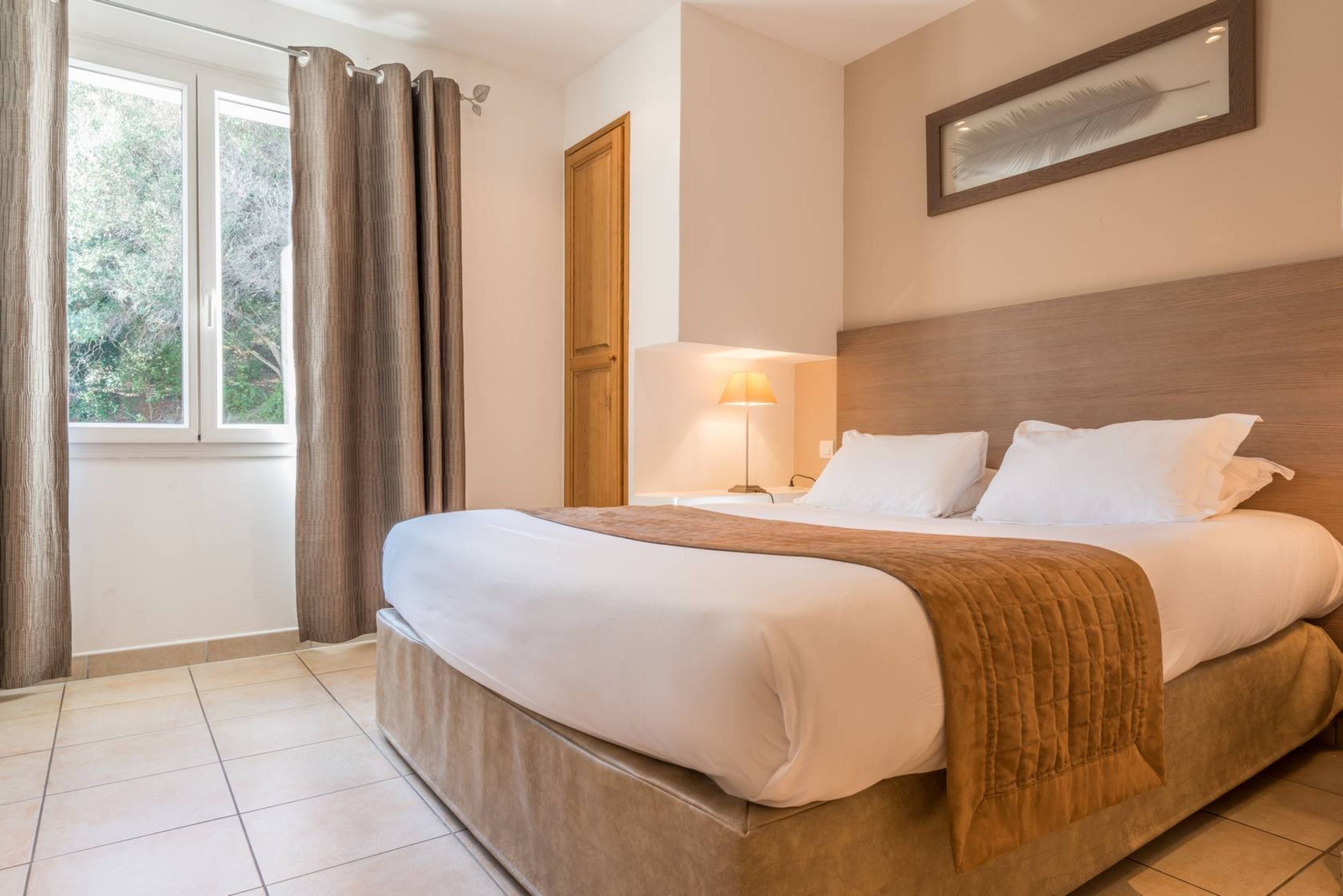 Una habitación de hotel con cama, mesita de noche, lámpara y ventana con cortinas.