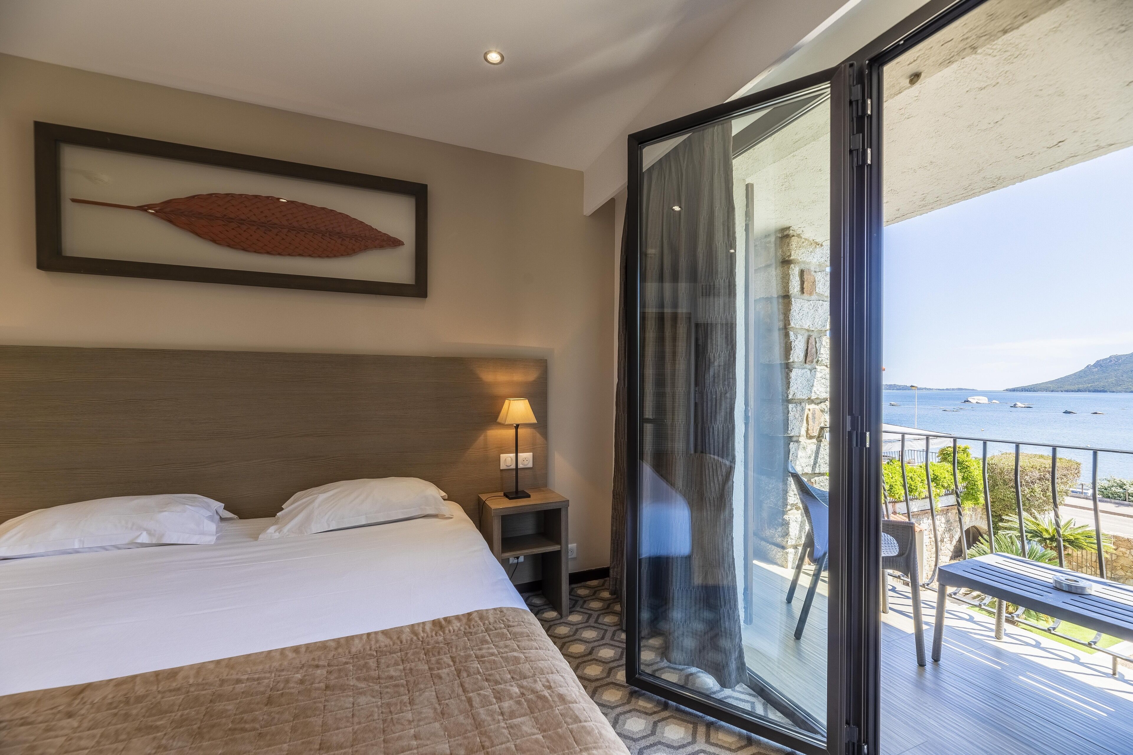 A hotel room with a bed, a nightstand, a framed leaf artwork, and a balcony with a view of the sea.