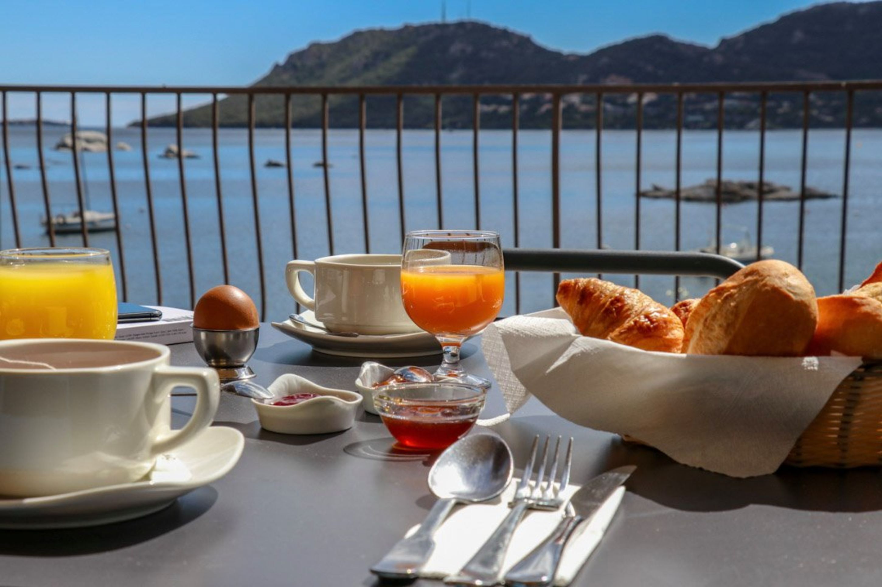 A breakfast setup with croissants, orange juice, and coffee on a table with a scenic view of a lake and mountains.
