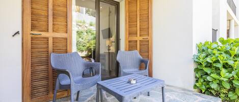 Un patio con dos sillones de mimbre, una mesita pequeña, una puerta de madera y un arbusto verde.