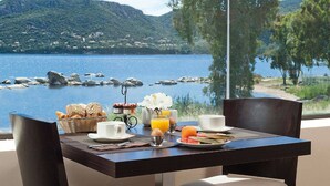 A breakfast table with a view of a lake and hills, including a basket of bread, a cup, a jug of orange juice, and a plate with a croissant and fruit. - Hôtel Shegara (Porto-Vecchio)