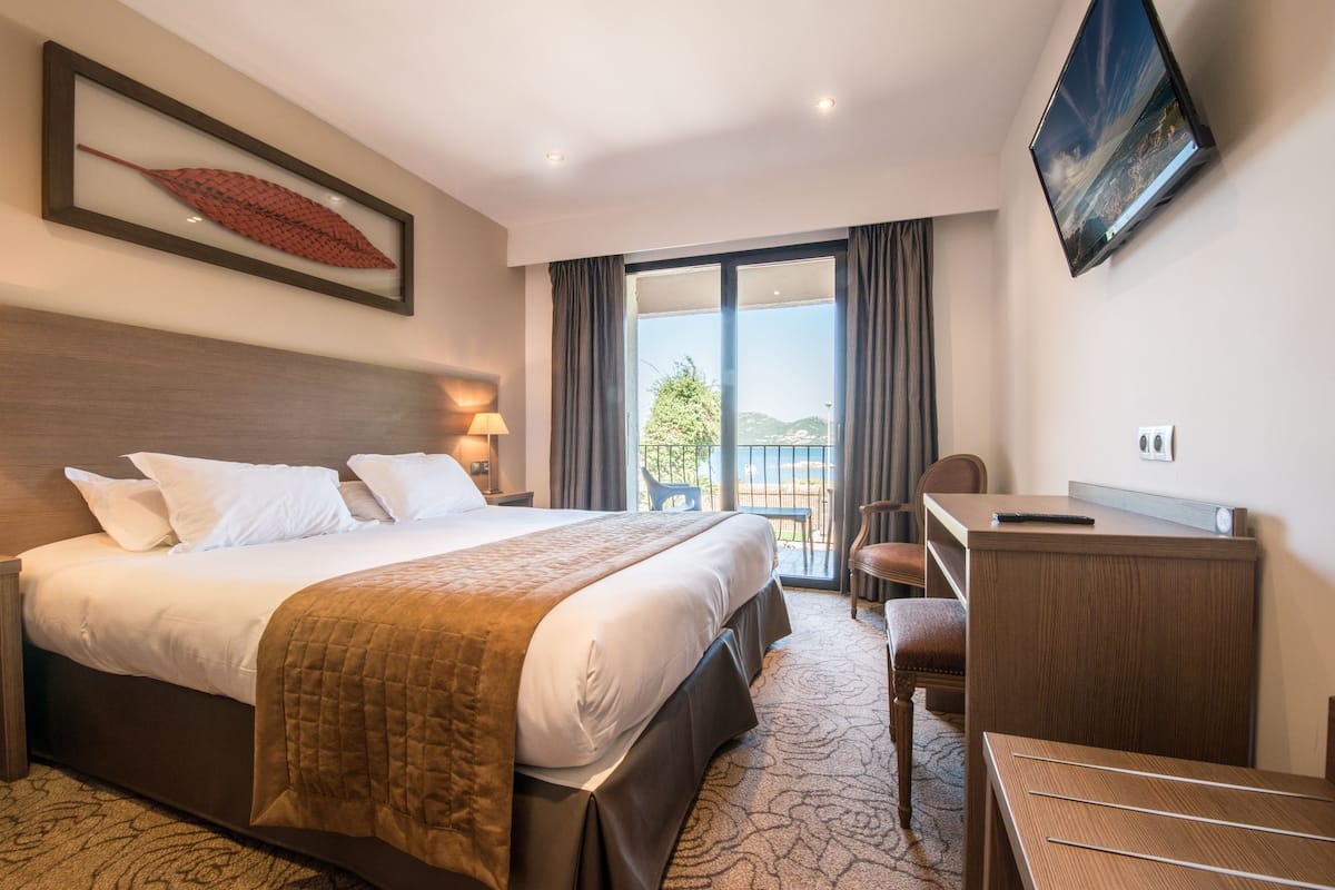 A hotel room with a large bed, a desk, a chair, a TV mounted on the wall, and a window with a view of the outdoors.