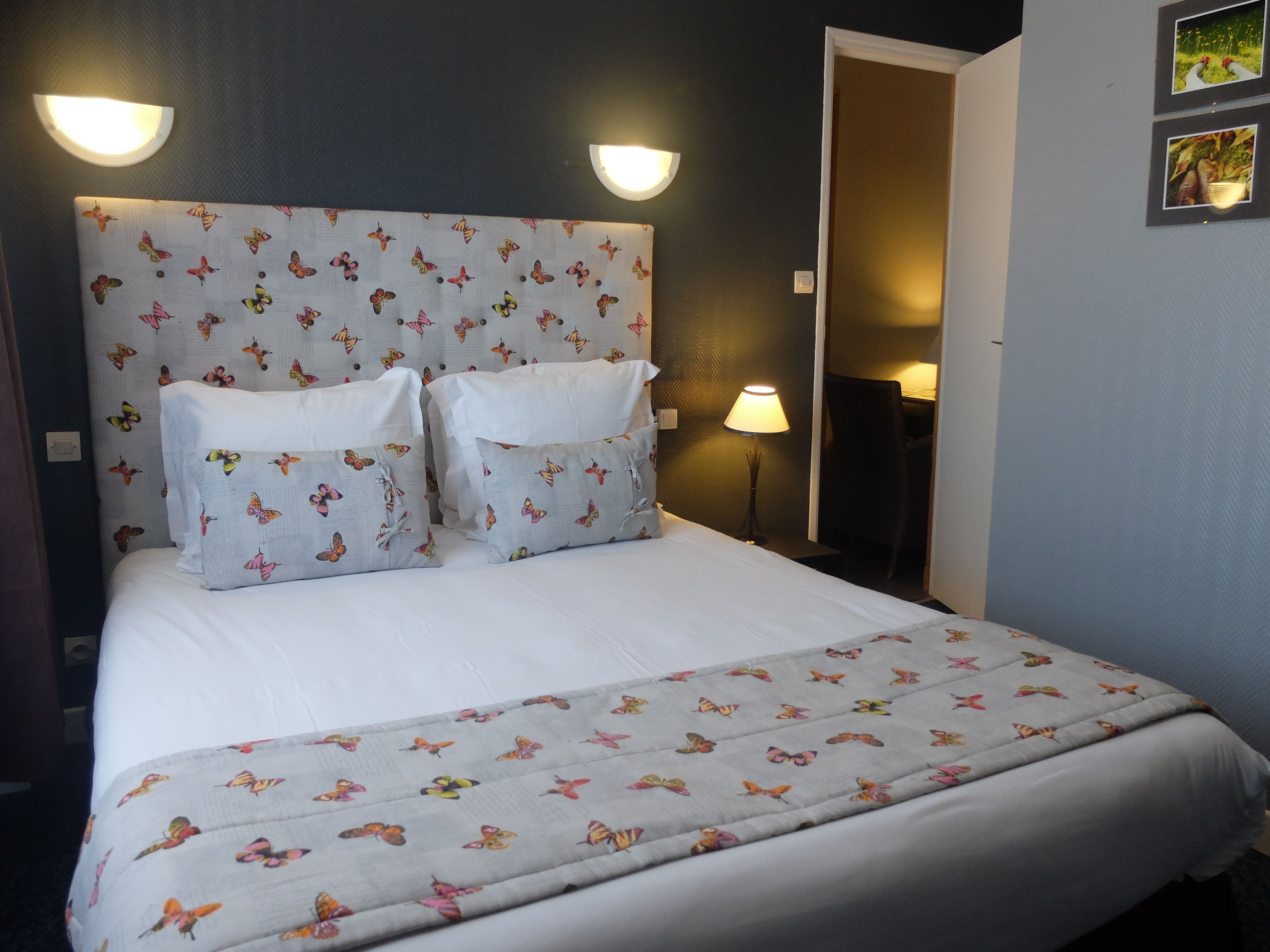Una cama bien hecha con una colcha y almohadas estampadas con motivos de mariposas, dos lámparas de pared y una puerta que da a otra habitación.