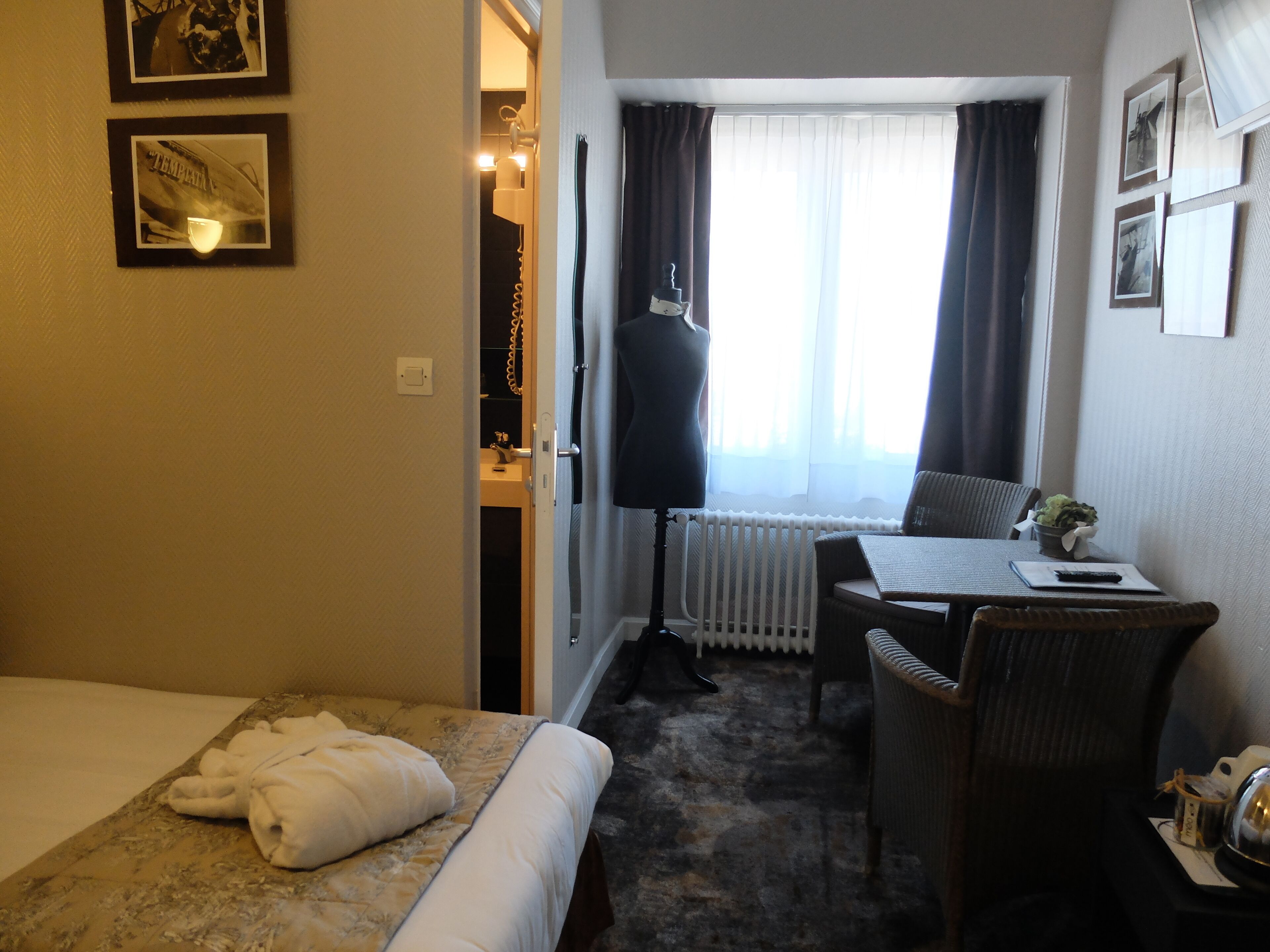 A hotel room with a bed, a desk with a chair, a radiator, and a window with curtains.