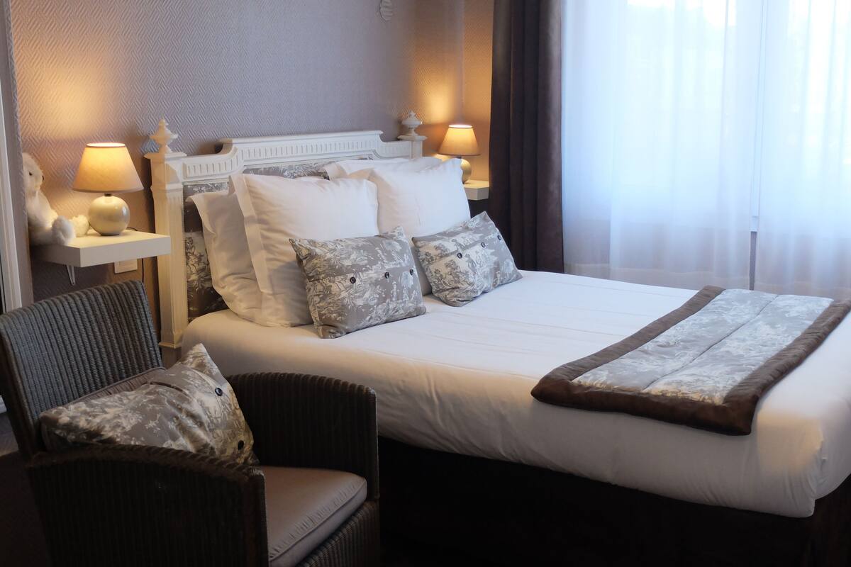 Hotelkamer met een groot bed, twee nachtkastjes met lampen, een stoel en uitzicht op het landschap door dunne gordijnen.