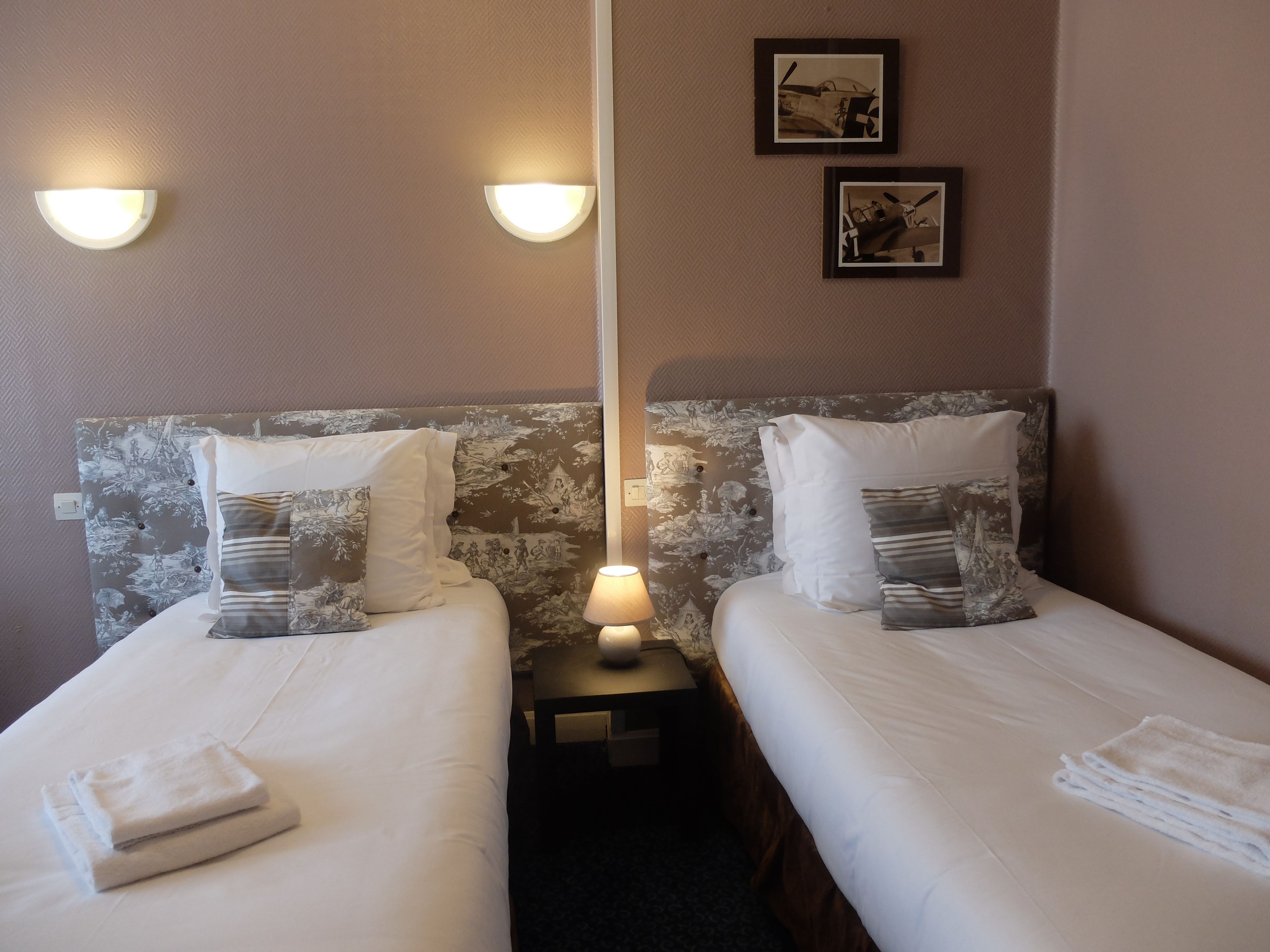 Une chambre d’hôtel avec deux lits, chacun doté de draps blancs et d’oreillers à motifs. Une table de chevet avec une lampe se trouve entre les deux lits. Les murs sont recouverts d’un papier peint texturé représentant un paysage naturel.