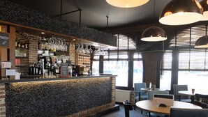 Una zona de bar con pared de piedra, estantes con botellas y vasos, y una barra con máquina de café. Hay mesas y sillas para sentarse.
