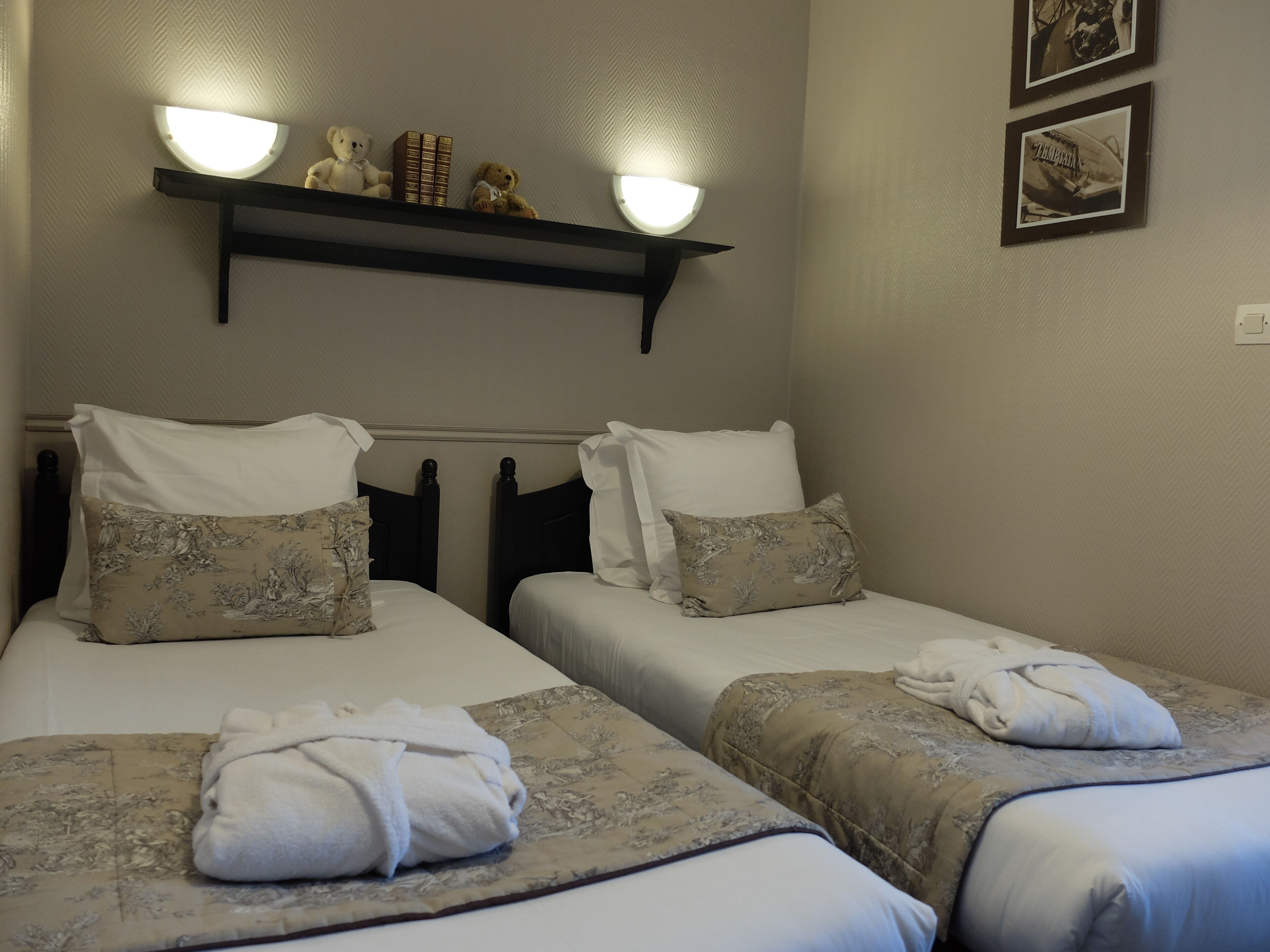 Quarto de hotel com duas camas, cada uma com almofada e toalha dobrada. Há uma estante acima das camas com objetos decorativos e uma luminária na parede.