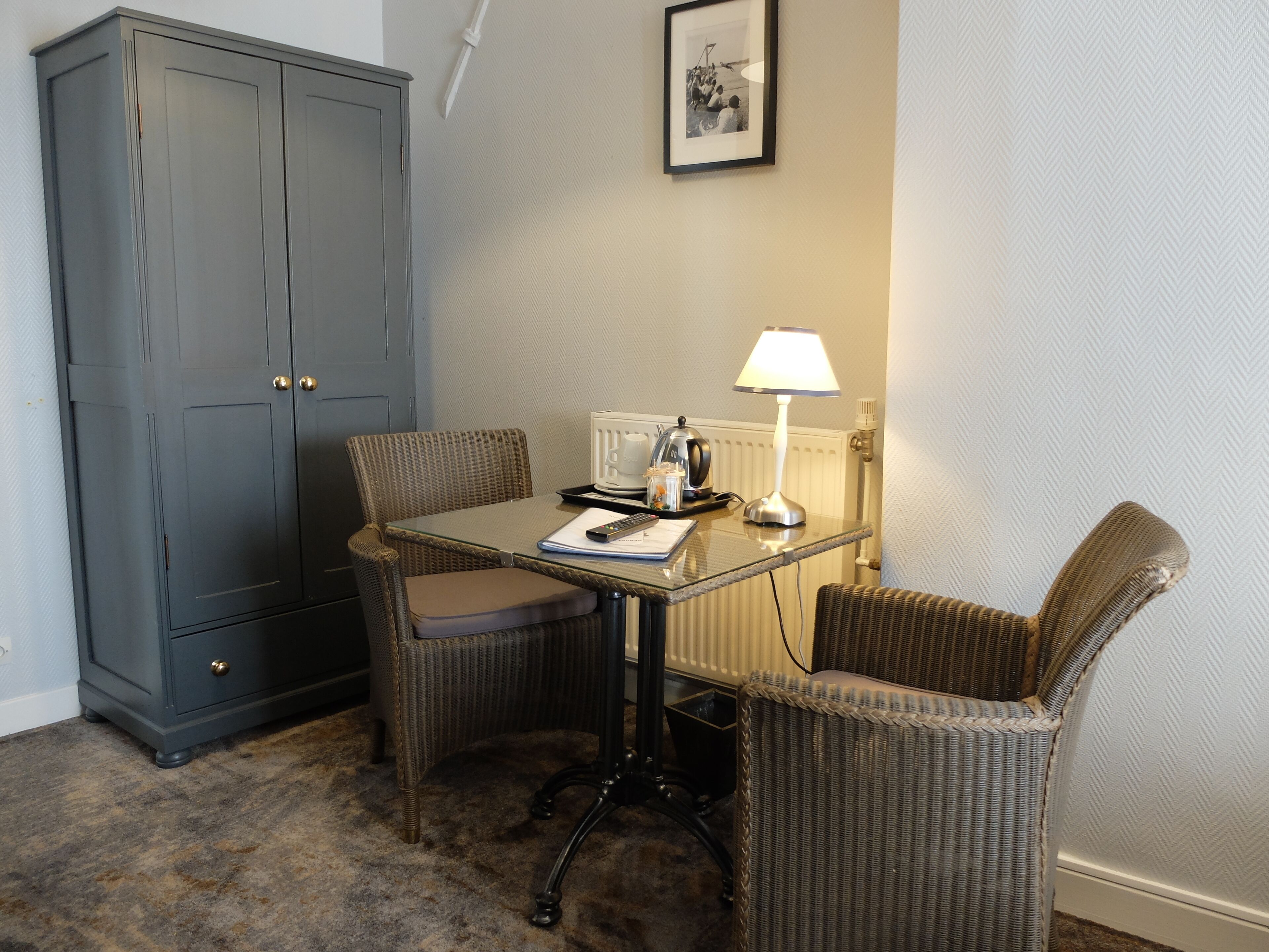 Een kleine kamer met een donkergrijze garderobe, een tafel met een lamp, een stoel en een ingelijst beeld aan de muur.