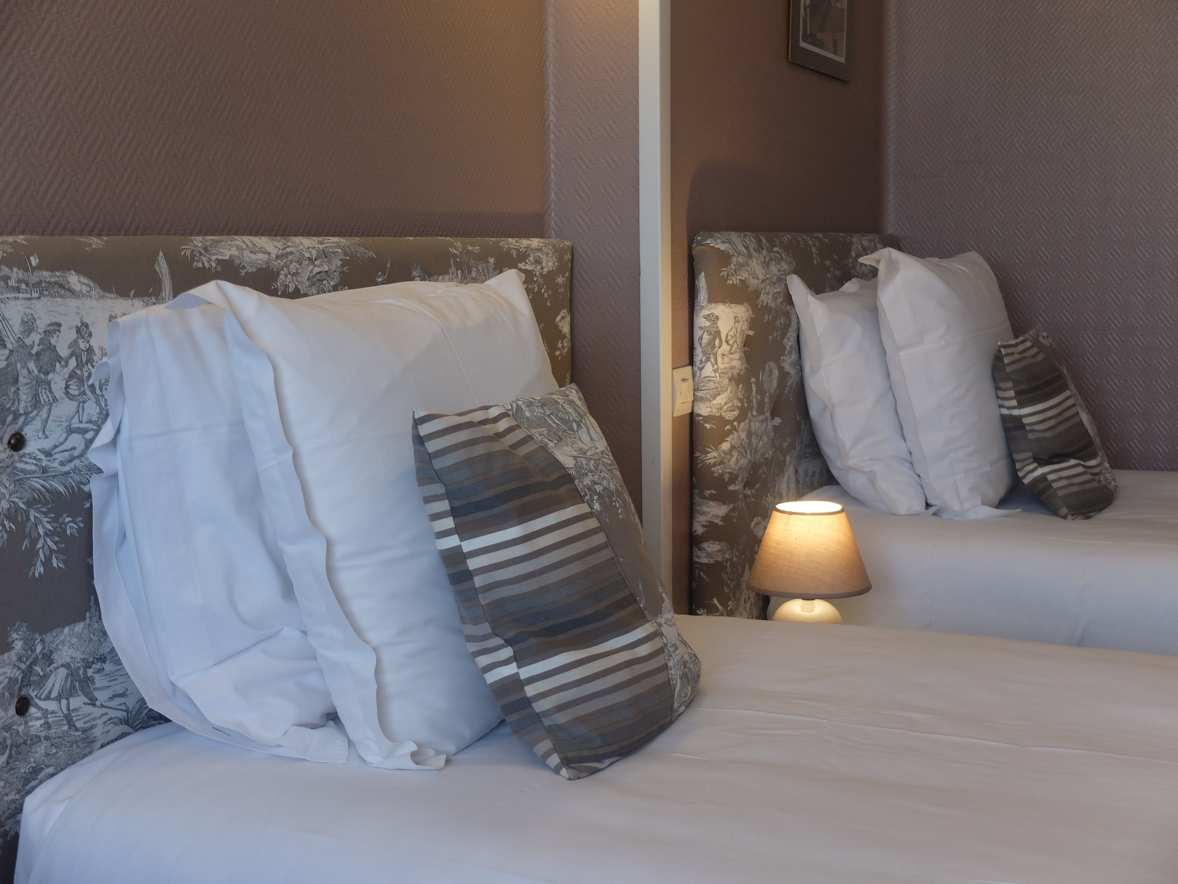 A hotel room with two beds, each with white bedding and striped pillows. A lamp is on the right side of the bed.