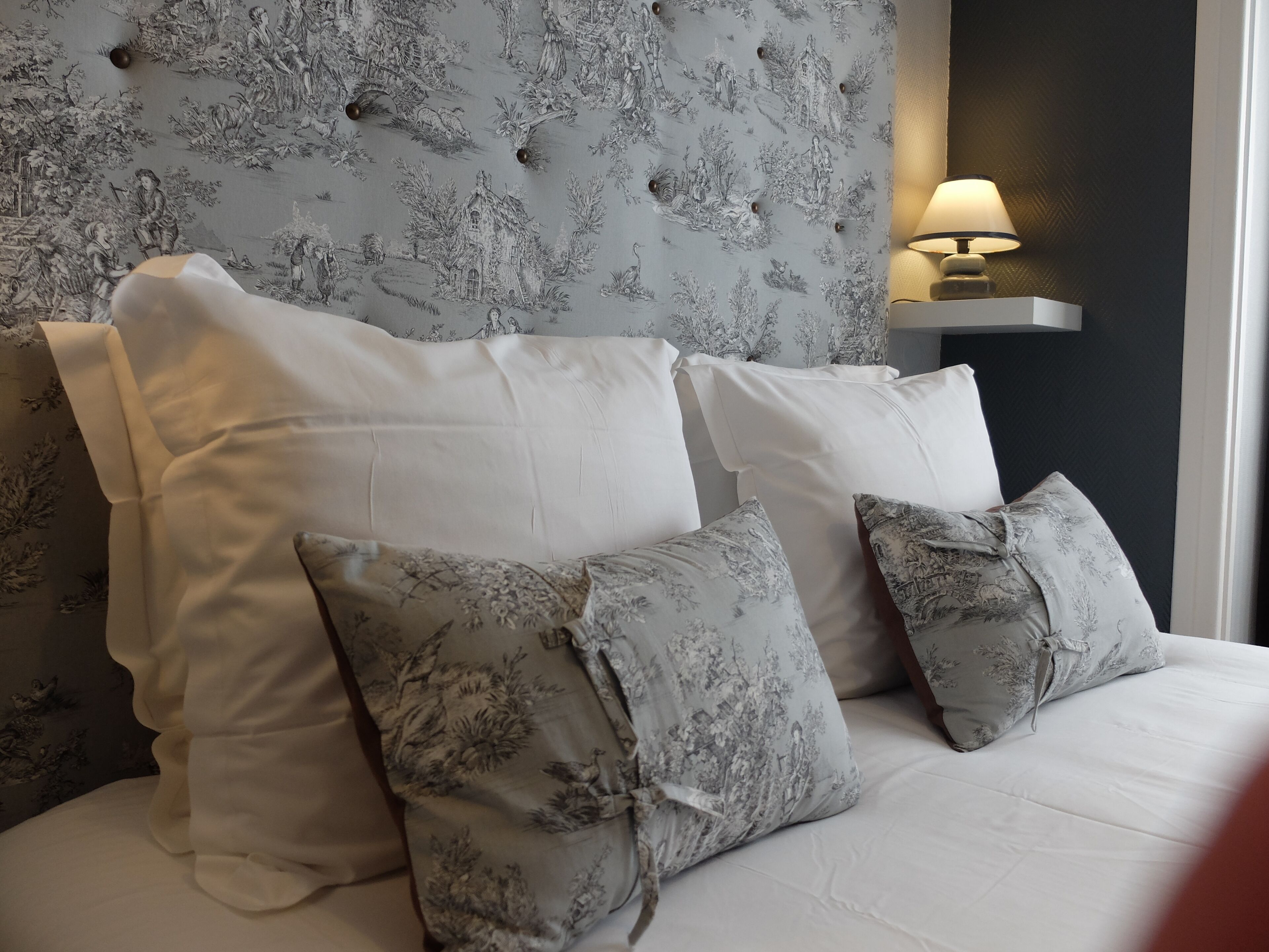 A bed with white bedding and grey floral patterned pillows, set against a wall with a grey floral wallpaper and a wall-mounted lamp.