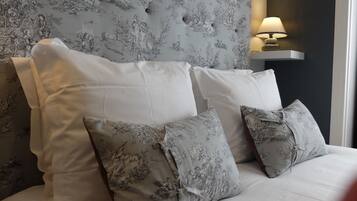 A bed with white bedding and grey floral patterned pillows, set against a wall with a grey floral wallpaper and a wall-mounted lamp.