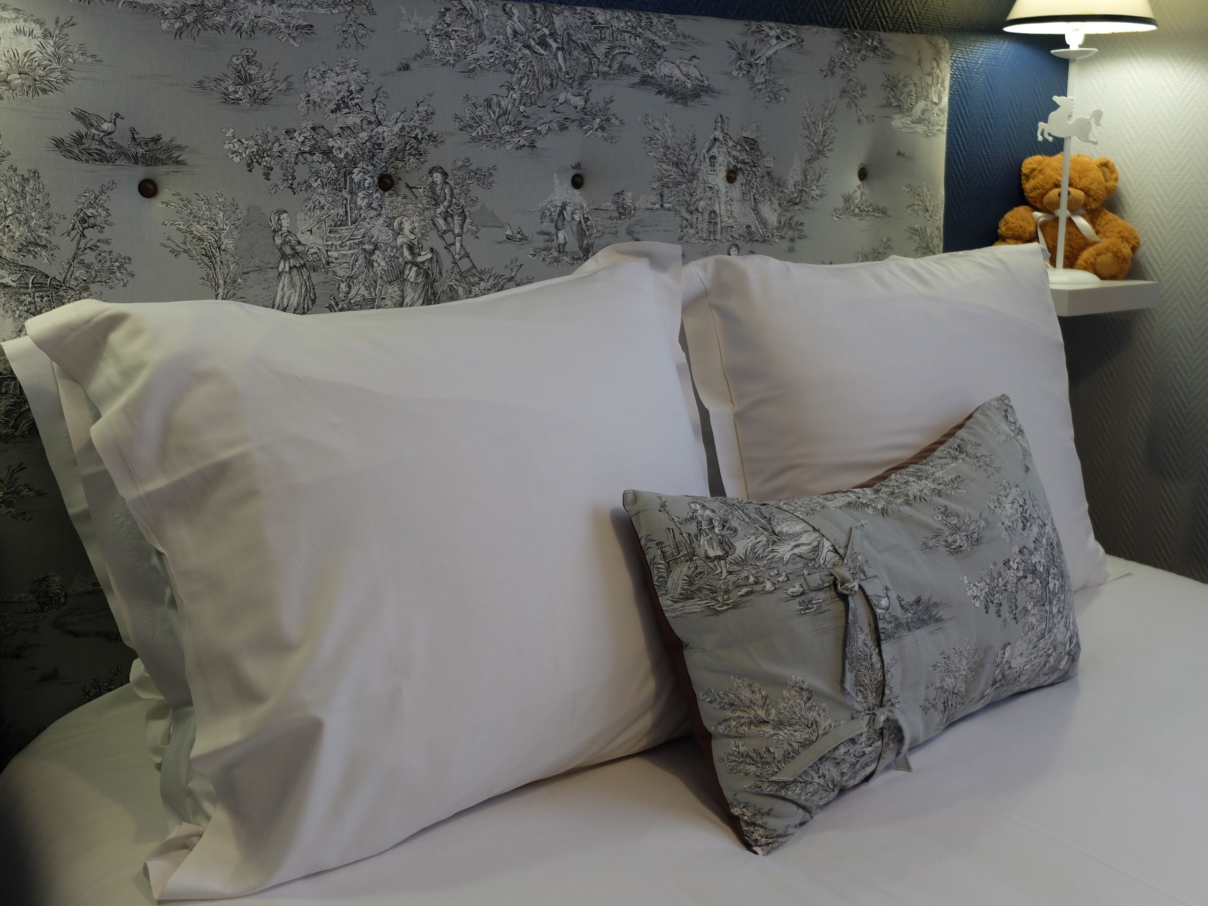 A bed with white and grey patterned pillows, a nightstand with a teddy bear, and a wall with a nature-themed wallpaper.