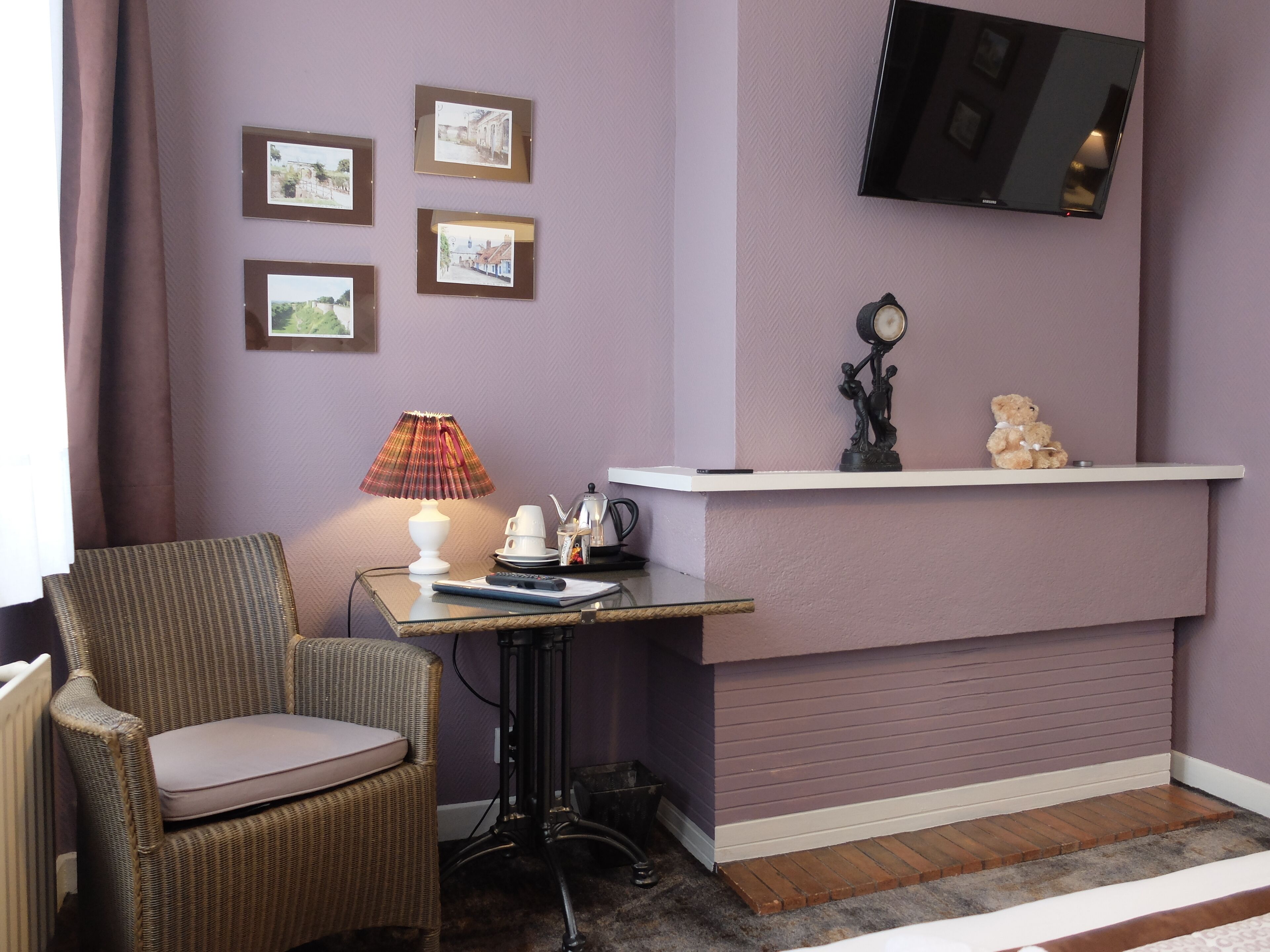 A corner room with a wicker chair, a small table with a lamp, a shelf with decorative items, and a mounted TV.