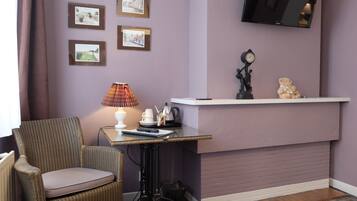 A corner room with a wicker chair, a small table with a lamp, a shelf with decorative items, and a mounted TV.