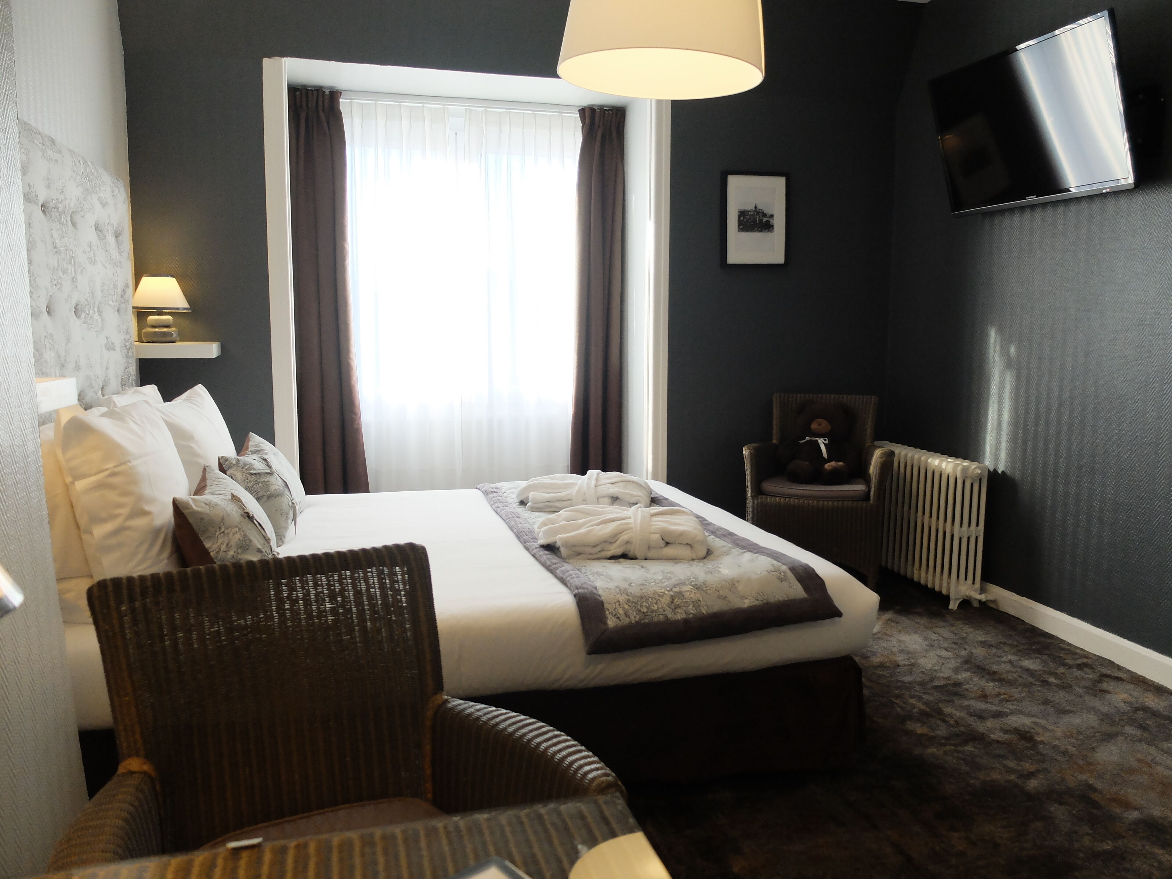 A hotel room with a bed, a chair, a bedside table with a lamp, a radiator, and a TV on the wall.
