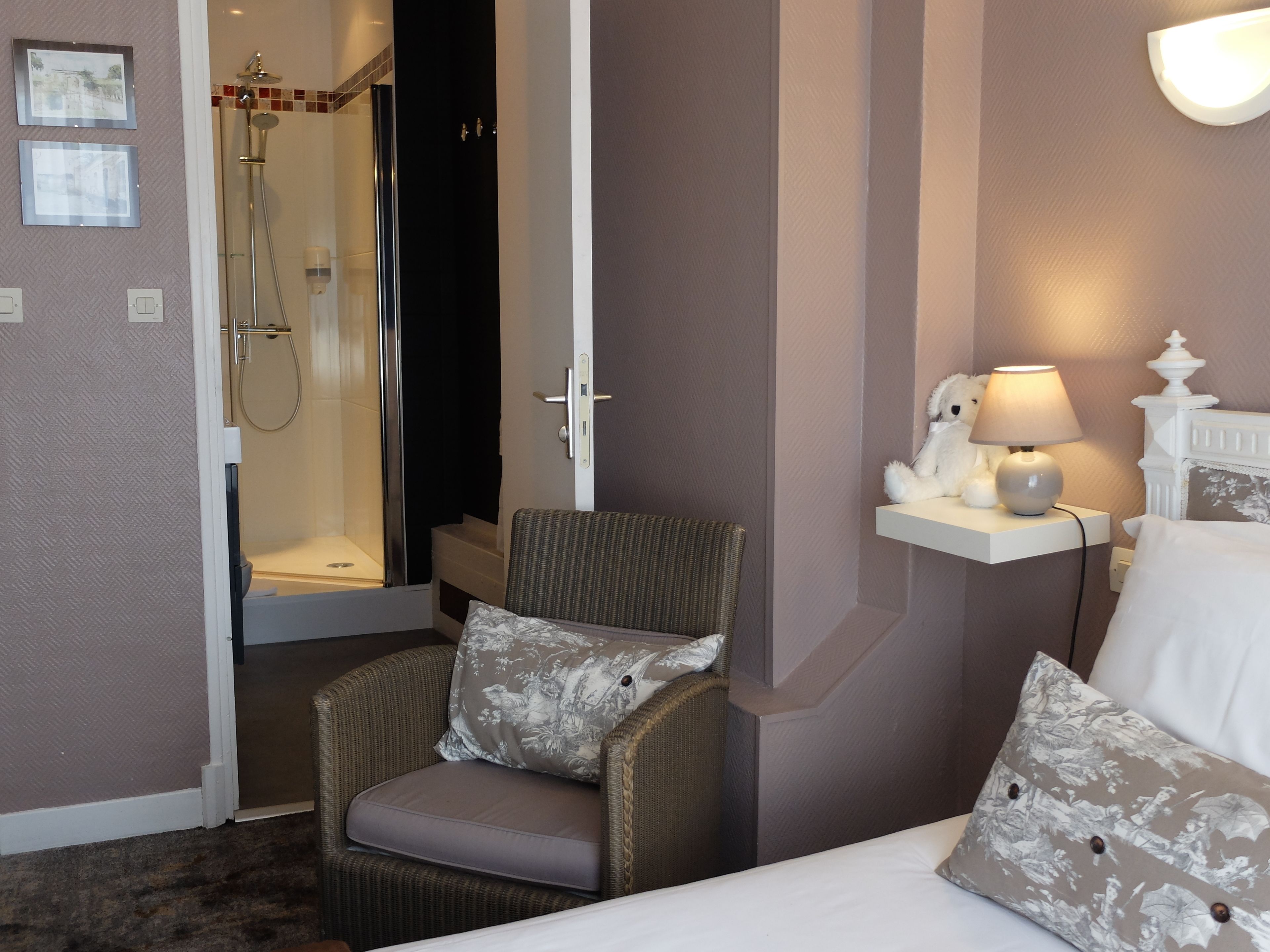 Habitación de hotel con cama, una silla, mesita de noche con lámpara, zona de ducha y estante en la pared con objetos decorativos.
