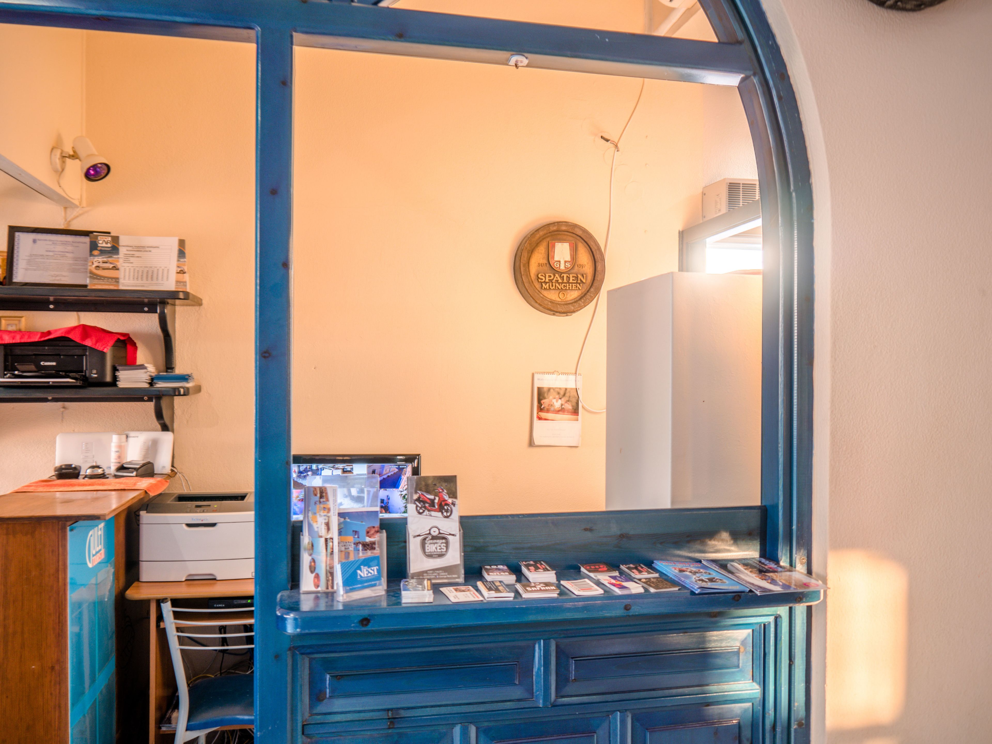 A room with a blue cabinet displaying books and magazines, a desk with a printer and computer, and a wall-mounted shelf with various items.