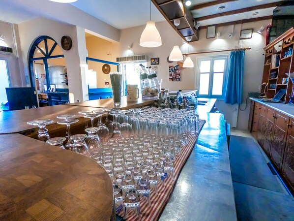 A bar area with multiple glasses and bottles, wooden furniture, and a blue curtain. - Princess Sissy Hotel (Ios)
